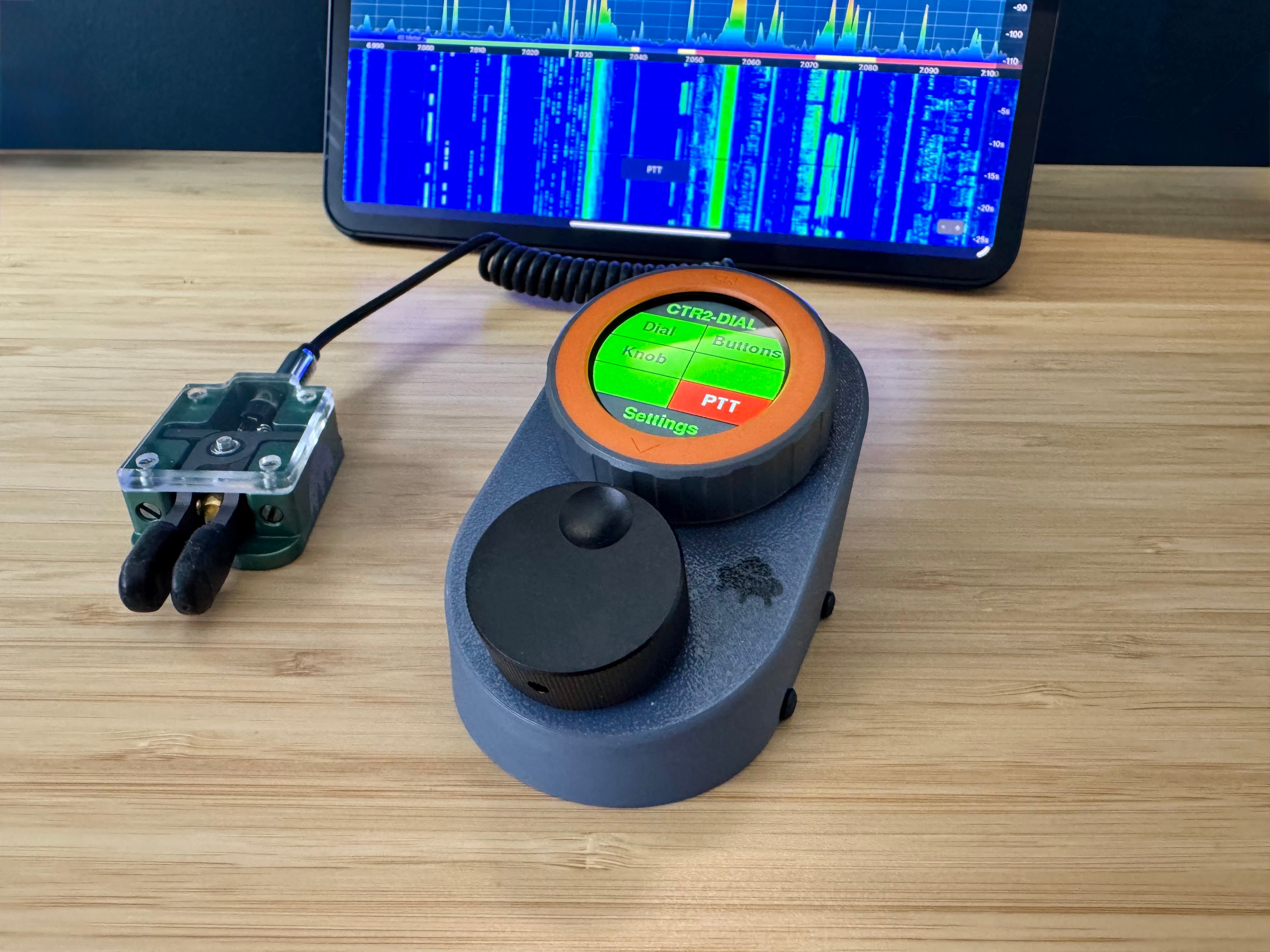 A CTR2-Dial in its grey case with the M5 Dial screen showing the PTT menu, connected to a paddle and an iPad running SmartSDR