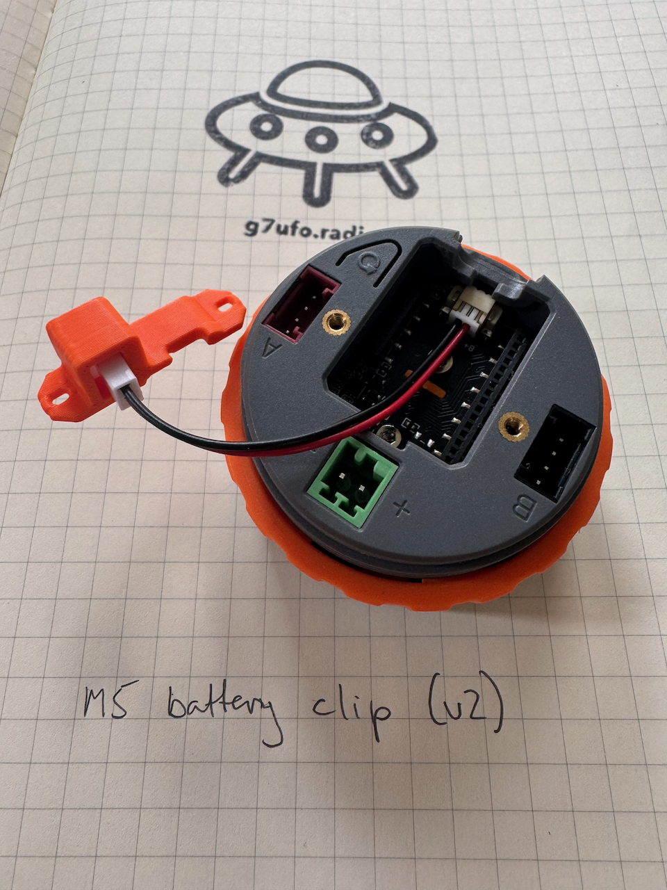The battery clip plugged into the M5 Dial The battery clip’s red and black cable plugged into the connector inside the M5 Dial with the STAMP S3A board removed, leading out to the orange battery adapter