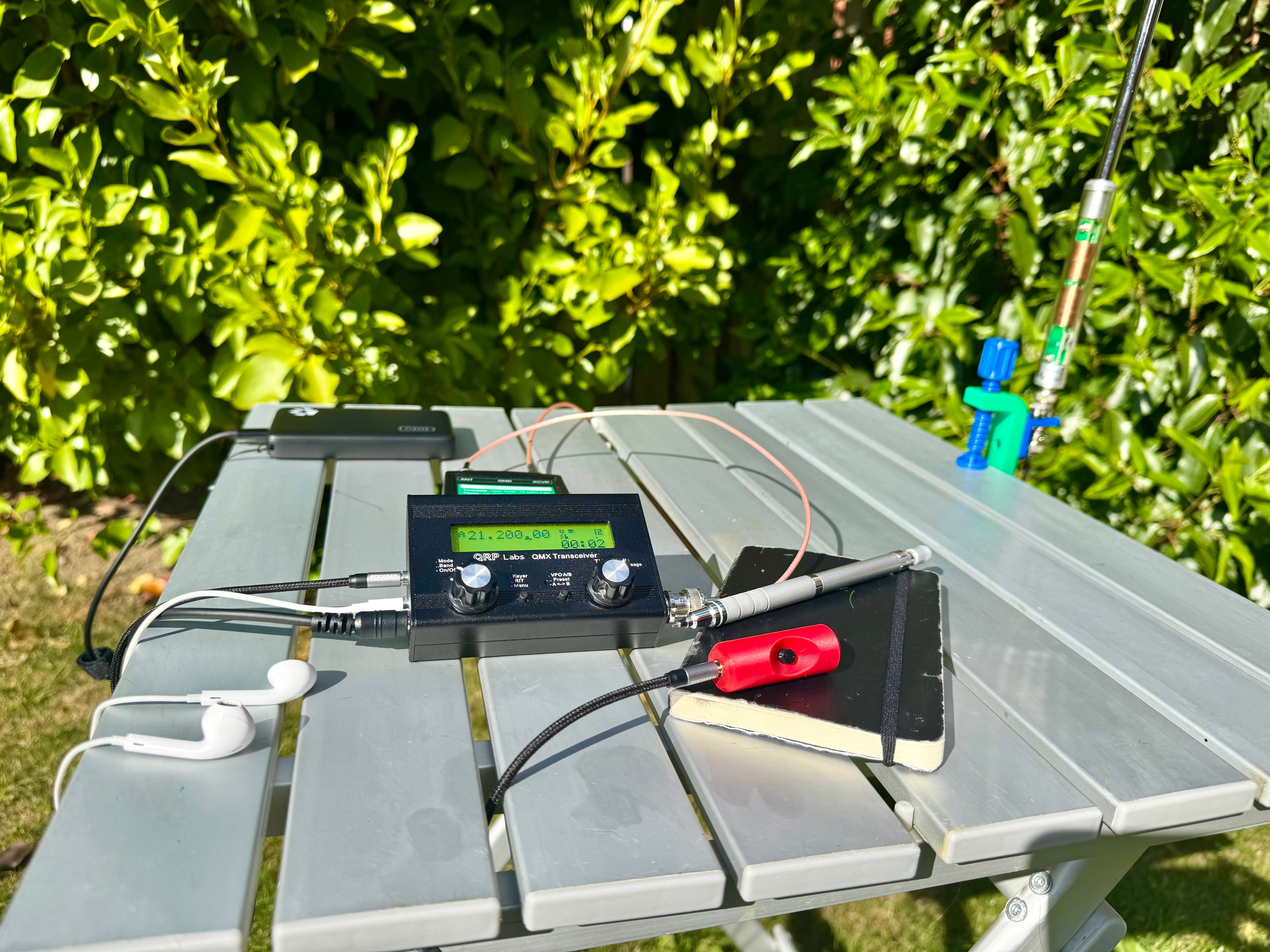 A QRP Labs QMX on a garden table, with a G7UFO turret microphone plugged in, a vertical antenna behind it and a notebook alongside